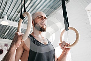 Muscular man holding gymnastic rings at light gym and looking away
