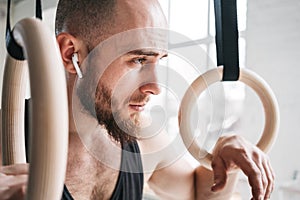 Strong bearded athlete with wireless headphone holding crosstraining rings at crossfit hall