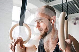 Strong bearded athlete with wireless headphone holding crosstraining rings at crossfit hall