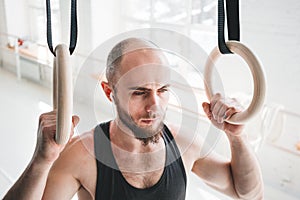 Strong bearded athlete with wireless headphone holding crosstraining rings at crossfit hall