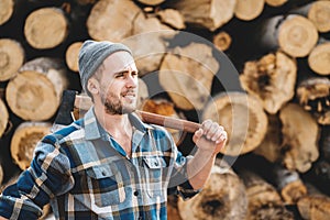 Strong bearded lumberjack wearing plaid shirt hold ax in hand on background of sawmill