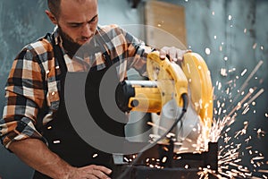 Strong bearded man working on electrical angular grinding machine in metalworking factory, sparks fly apart.