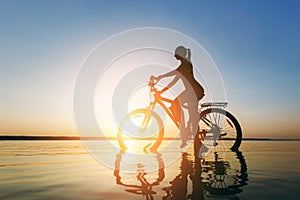 A strong blonde woman in a colorful suit sits on the bicycle in the water at sunset on a warm summer day. Fitness concept. Sky bac
