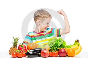 Strong boy shows biceps at the table with a pile of fresh vegetables and fruits isolated