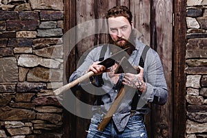 Strong brutal man with a beard and tattoos on his hands dressed in stylish casual clothes stands with two axes in his
