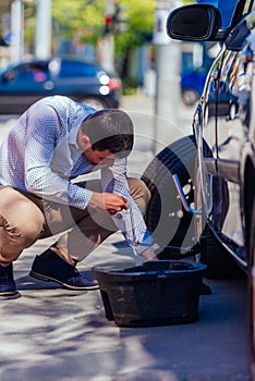 A strong businessman is bending his knee while trying to change a flattie on his car with his lug wrench