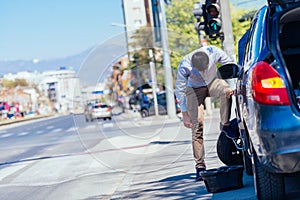 A strong businessman is bending his knee while trying to change a flattie on his car with his lug wrench