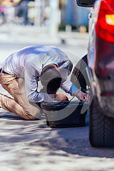 A strong businessman is bending his knee while trying to change a flattie on his car with his lug wrench