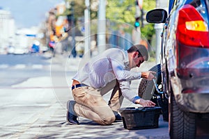 A strong businessman is bending his knee while trying to change a flattie on his car with his lug wrench