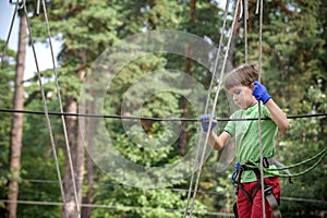 Strong excited young boy playing outdoors in rope park. Caucasian child dressed in casual clothes and sneakers at warm sunny day.