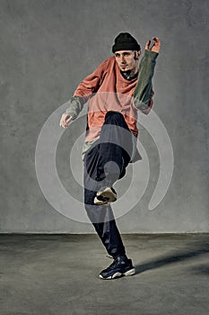 Strong fellow with tattooed face, beard. Dressed in hat, colorful jumper, black pants and sneakers. Dancing, gray