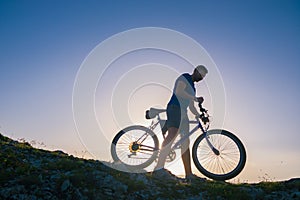 Strong fit male mountain biker performing stunts on rocky terrain on a sunset while wearing a blue shirt and riding a blue bike