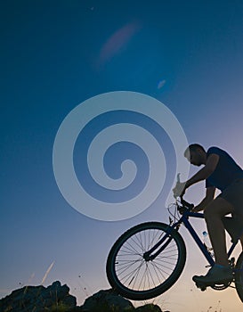Strong fit male mountain biker performing stunts on rocky terrain on a sunset while wearing a blue shirt and riding a blue bike