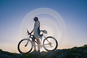 Strong fit male mountain biker performing stunts on rocky terrain on a sunset while wearing a blue shirt and riding a blue bike