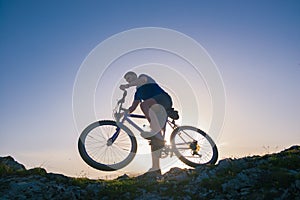 Strong fit male mountain biker performing stunts on rocky terrain on a sunset while wearing a blue shirt and riding a blue bike