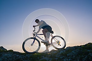 Strong fit male mountain biker performing stunts on rocky terrain on a sunset while wearing a blue shirt and riding a blue bike