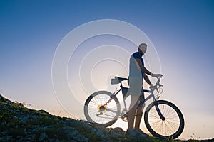 Strong fit male mountain biker performing stunts on rocky terrain on a sunset while wearing a blue shirt and riding a blue bike