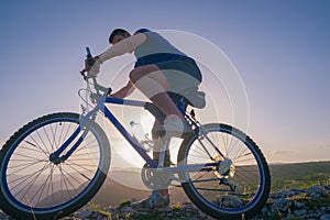 Strong fit male mountain biker performing stunts on rocky terrain on a sunset while wearing a blue shirt and riding a blue bike