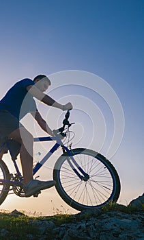 Strong fit male mountain biker performing stunts on rocky terrain on a sunset while wearing a blue shirt and riding a blue bike