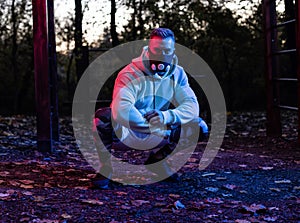 Strong good looking young man with workout fitness mask in squatting position in the park taking a break from workout as dusk sets
