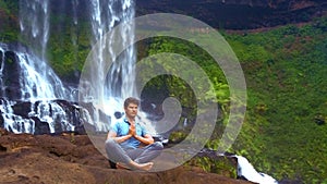 Strong guy sits in pranayama yoga pose on rock at waterfall