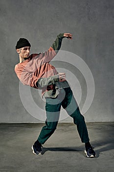 Strong guy with tattooed body, earrings, beard. Dressed in hat, casual clothes and black sneakers. Dancing on gray