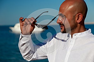 Strong handsome beautiful man in black fashion sunglasses, white shirt enjoys vacation trip at sea takes suntan at sunny summer