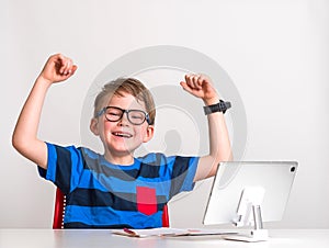 Strong little blonde kid in glasses has online lesson. 7, 8 years old in striped blue shirt doing homework. Smiling boy