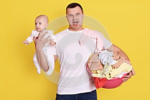 Strong male in white casual t shirt holding her new born baby and basin in hands, doing laundry, carries dirty clothing, standing