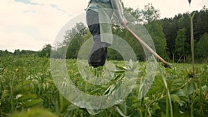 Strong man 60-65 years old with a scythe mows green grass on the field.