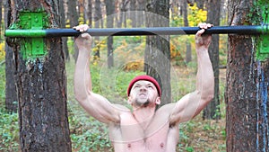 Strong man doing pull ups on horizontal bar with crazy expression on face. Male sportsman exercising at nature. Young