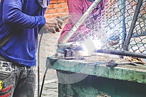 Strong man is a welder in a blue T-shirt,and welders leathers, a metal product is welded with a welding machine in the outdoor photo