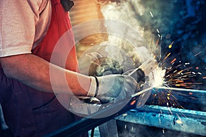 Strong man is a welder in T-shirt in a construction gloves, a metal product is welded with a welding machine in the garage