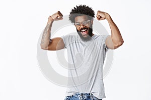Strong and masculine good-looking happy african american male with beard in glasses showing biceps as raising hands in