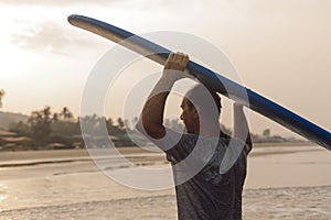 Strong middle aged man carrying blue long surfing board on head coming out of sea and looking at beach on sunset.