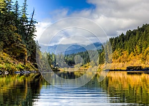 Strong mossy boulders line the edges of the lower Rogue River in late October with blue sky and clouds in distance