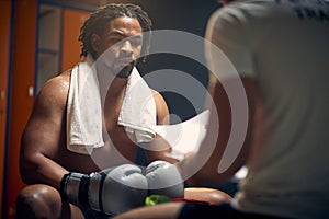 Strong muscular boxer full of sweat after boxing workout resting, sitting in dressing room with his coach