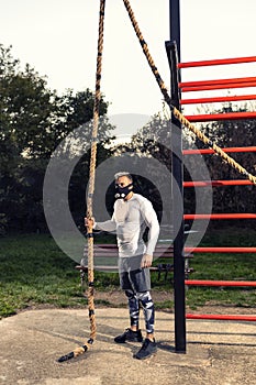 Strong muscular man wearing a workout breathing mask getting ready to climb a rope outdoors