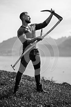 Strong muscular power sexy farmer using scythe. Farmer with a scythe on green grass field. Guy cut grass. Rural farmer