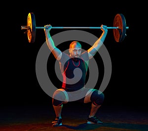 Strong, muscular, young athlete, bodybuilder doing squats and lifting barbell, heavy weights against blue background in