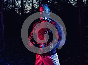 Strong muscular young man with weightlifting chains around his body and a fitness mask, working out in the park as evening