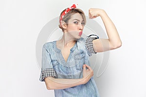 Strong powerful woman in blue denim shirt raised her arm, showing biceps, keeps lips pout.