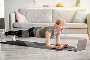 Strong senior woman standing in elbow plank on yoga mat, working out to online sports video at home