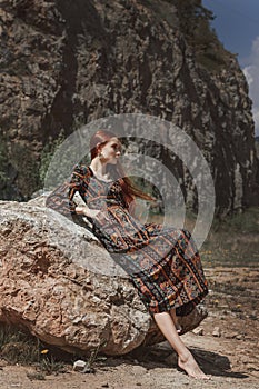 Strong-willed free strong red-haired woman in an ethnic dress near a big stone. A symbol of indomitability
