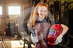 Strong and worthy pretty redhead ginher woman wearing protection helmet and leather apron with gloves holding welding