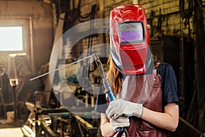 Strong and worthy pretty redhead ginher woman wearing protection helmet and leather apron with gloves holding welding