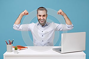 Strong young bearded man in light shirt sit, work at desk with pc laptop isolated on pastel blue background. Achievement