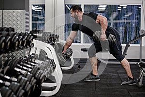 Strong young bearded shouting man pull dumbbell in one hand standing in slope in sport gym