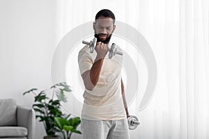 Strong young black male with beard in white sportswear raises dumbbell in room interior on window background