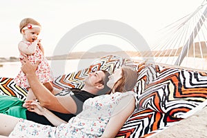 Strong young father holding his baby daughter in hands with happy hipster mother, smiling family relaxing in a hammock on a beach